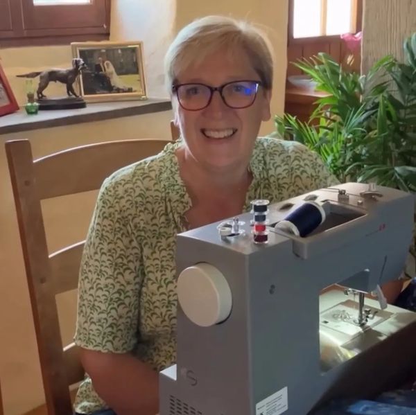 A smiling woman sits behind a sewing machine at a wooden table with fabric and scissors.