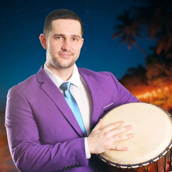 Man in purple suit holding a drum at night with palm trees in the background.