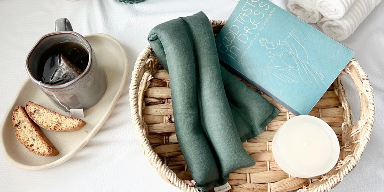 A cozy setup with a emerald green linen microwavable neck wrap, hot mug of tea, biscotti, a vintage fashion book, and a scented candle on a woven tray