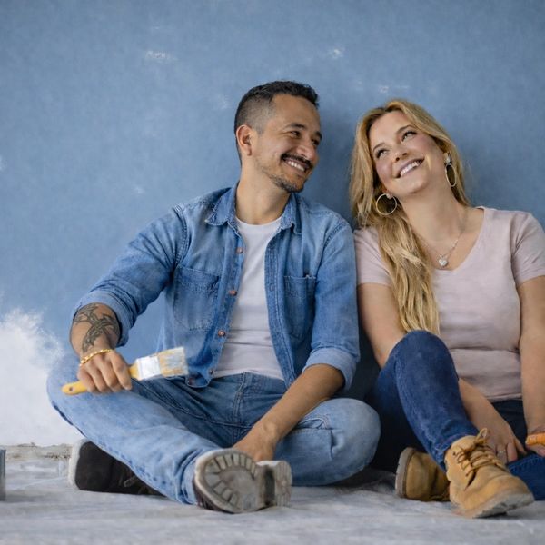 Couple sitting on the floor, smiling while holding paintbrushes against a blue wall.