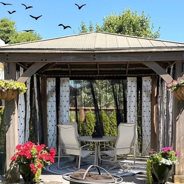 Cozy outdoor gazebo with seating, flowers, and birds flying overhead on a sunny day.