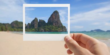 Hand holding photo of limestone cliffs over tropical beach in Krabi, Thailand.