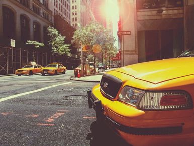 Yellow taxis line a sunny city street with buildings and trees.