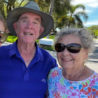 Elderly couple smiling outdoors on a sunny day with palm trees in the background.