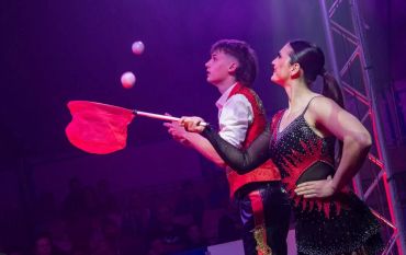 Two performers juggling with red clubs under purple lighting.