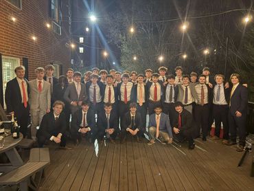 Large group of young men dressed formally, posing outdoors at night with string lights overhead.