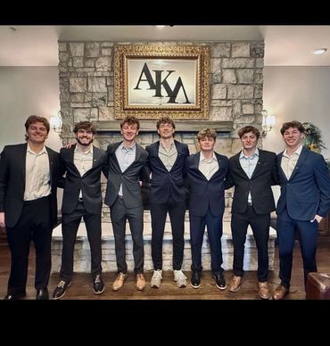 Seven young men in suits standing in front of a stone fireplace with a framed "AKA" sign.