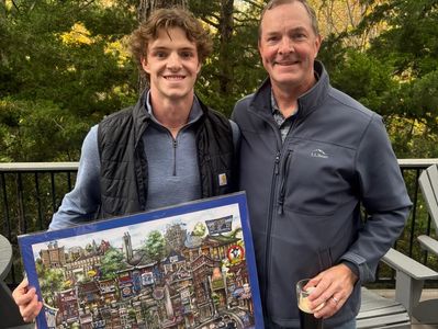 Two men smiling outdoors holding a colorful framed artwork.