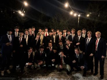 A large group of young men dressed in suits posing outdoors at night under string lights.
