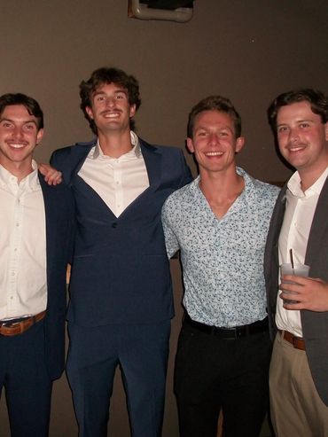 Four young men dressed smartly, smiling and posing together indoors.