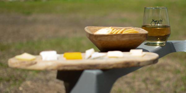 Wooden board with cheese slices, crackers in a bowl, and a glass of drink outdoors.