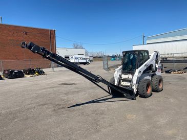 A skid steer loader equipped with an extended conveyor attachment outdoors.