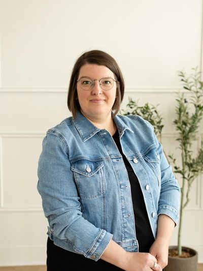 Woman in a denim jacket posing indoors with a plant in the background.