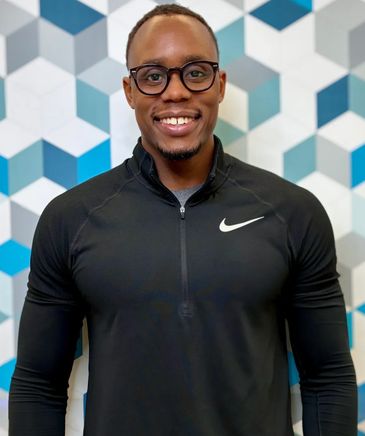 Smiling man in black Nike top with glasses stands before geometric patterned wall.