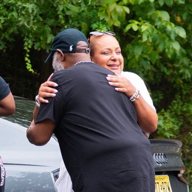 Two people warmly hugging outdoors near cars and greenery.