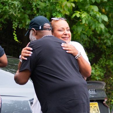 Two people warmly hugging outdoors near cars and greenery.