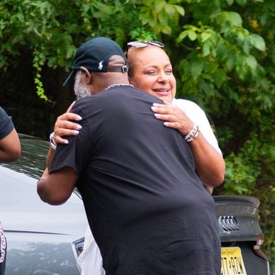 Two people warmly hugging outdoors near cars and greenery.