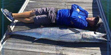 Man in blue lies next to a large fish on a dock.