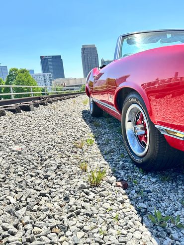 Red classic car parked on gravel near train tracks.