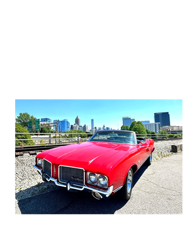 Red vintage convertible car parked near railway tracks with city skyline.