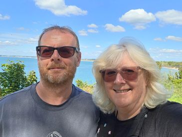 A smiling couple wearing sunglasses outdoors with a lake and blue sky behind them.