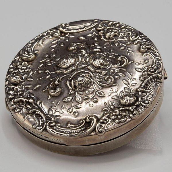 Ornate round silver compact with floral and scrollwork design.