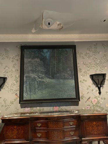 Damaged ceiling above a forest painting and wooden dresser.