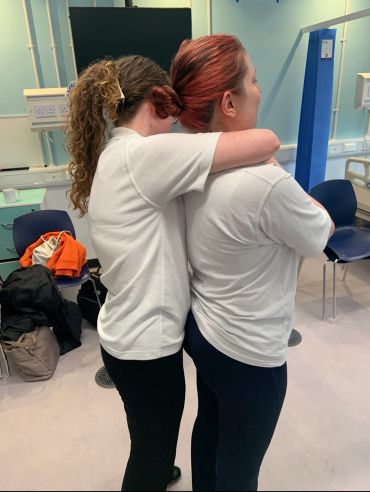 Two women in white shirts hugging from behind in a room.