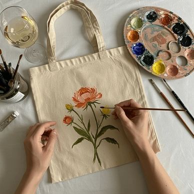 Person painting a floral design on a beige tote bag with watercolors.