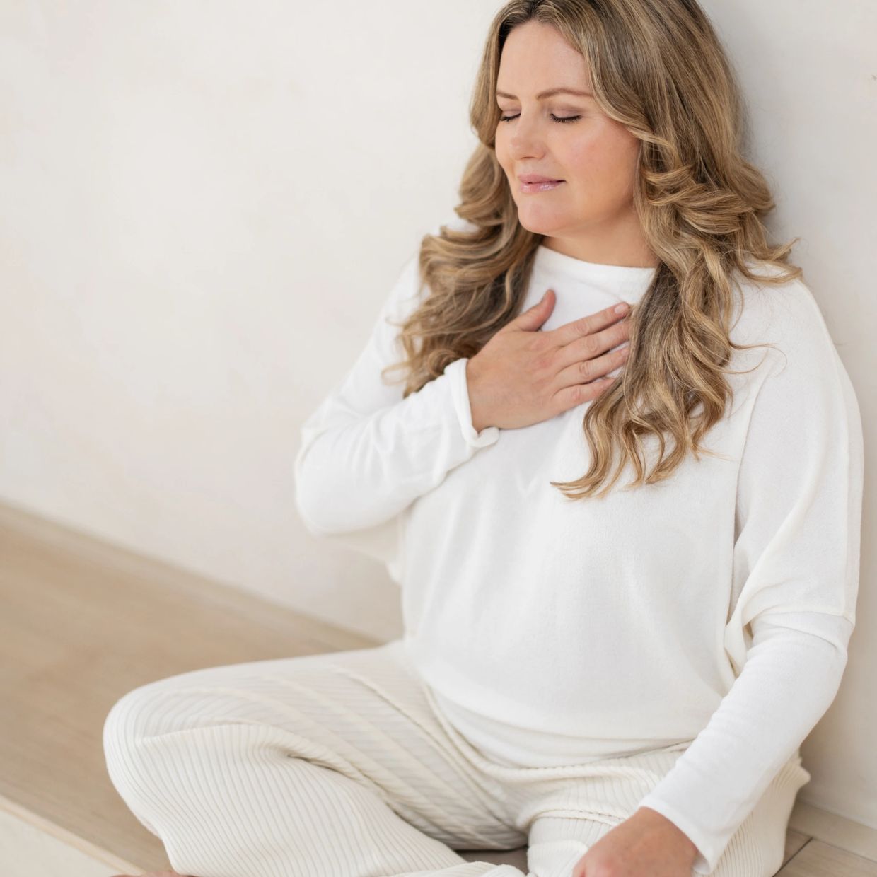 A woman in white sitting calmly with her hand on her chest, eyes closed.