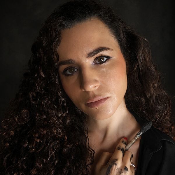 Woman with curly hair holding a paintbrush, with paint on her fingers.