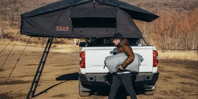 Man carrying a rolled-up sleeping bag near a rooftop tent on a white truck.