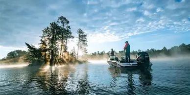 Fisherman on a boat during misty sunrise on a lake near trees.