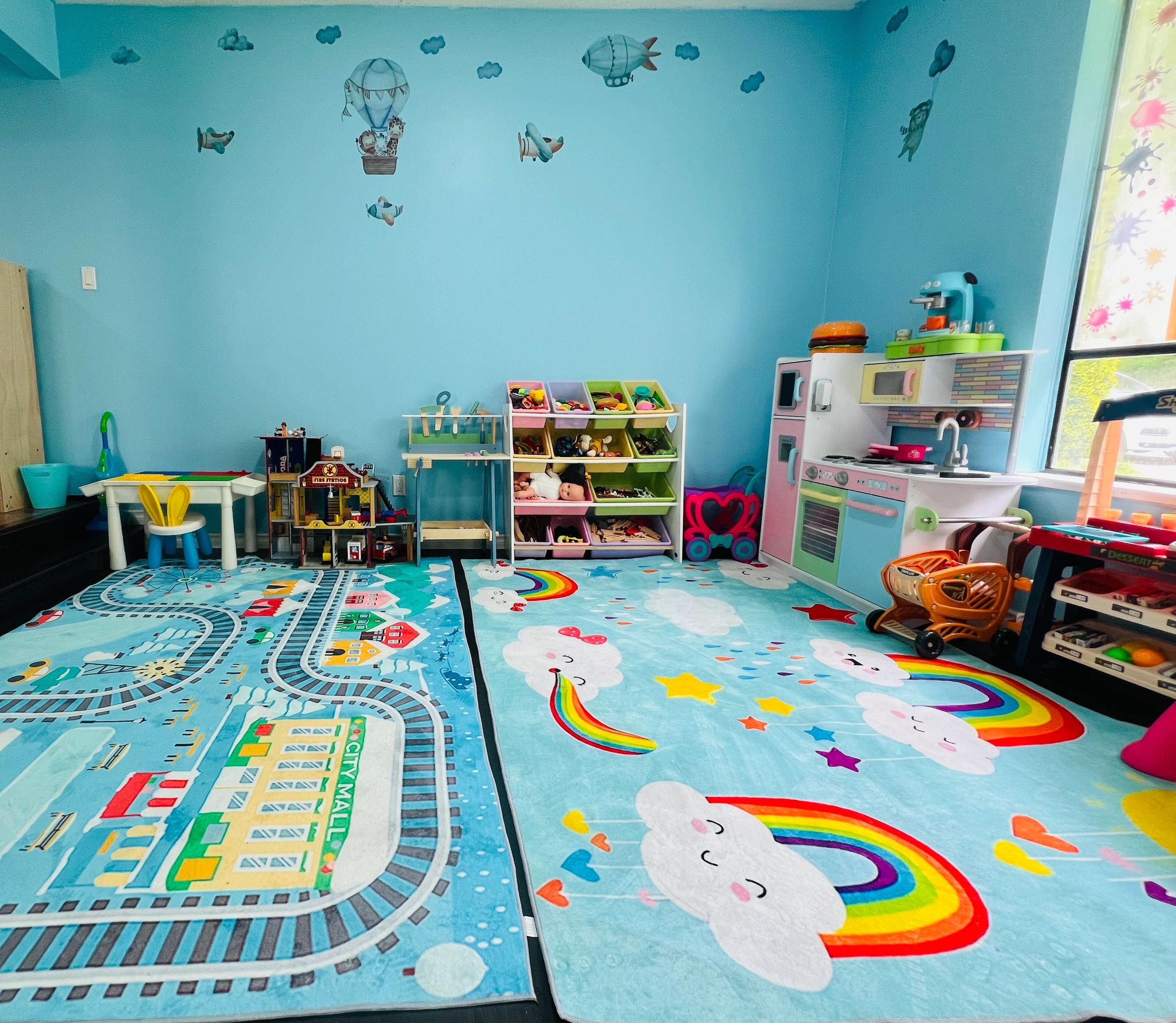 Colorful kids' playroom with toys, rugs, and a mini kitchen setup under blue walls with cloud and airplane decals.