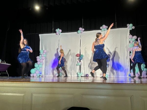 Four dancers in blue sequined dresses perform on a decorated stage.