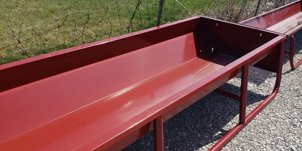 Long red metal livestock feeder outdoors on gravel near a fence.