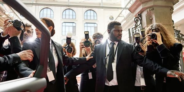 Security guards protect a VIP from photographers in a crowded area.
