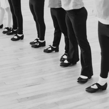 Line of dancers wearing black shoes and leggings on wooden floor.