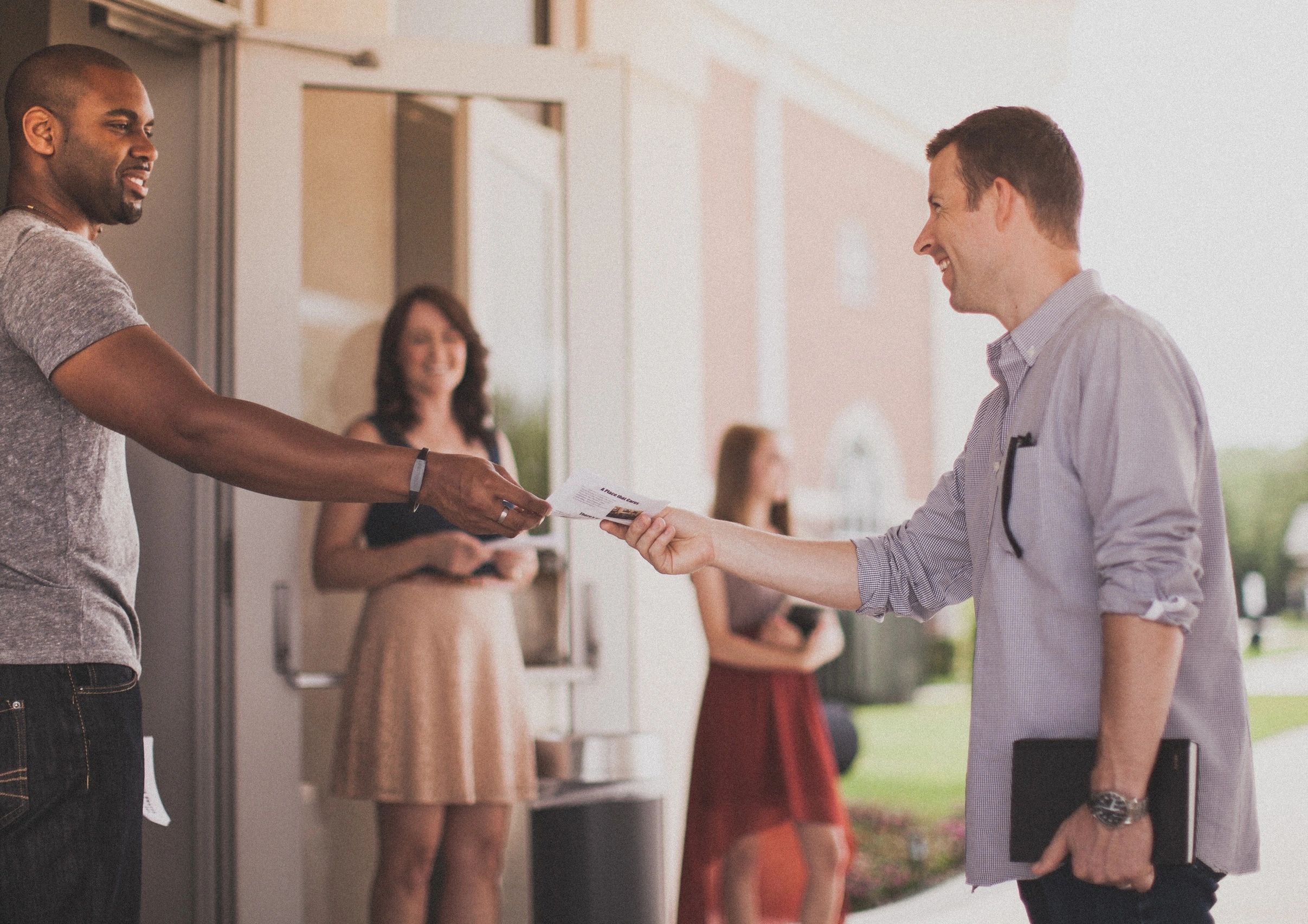 Two men exchanging a paper outdoors with two women smiling in the background.