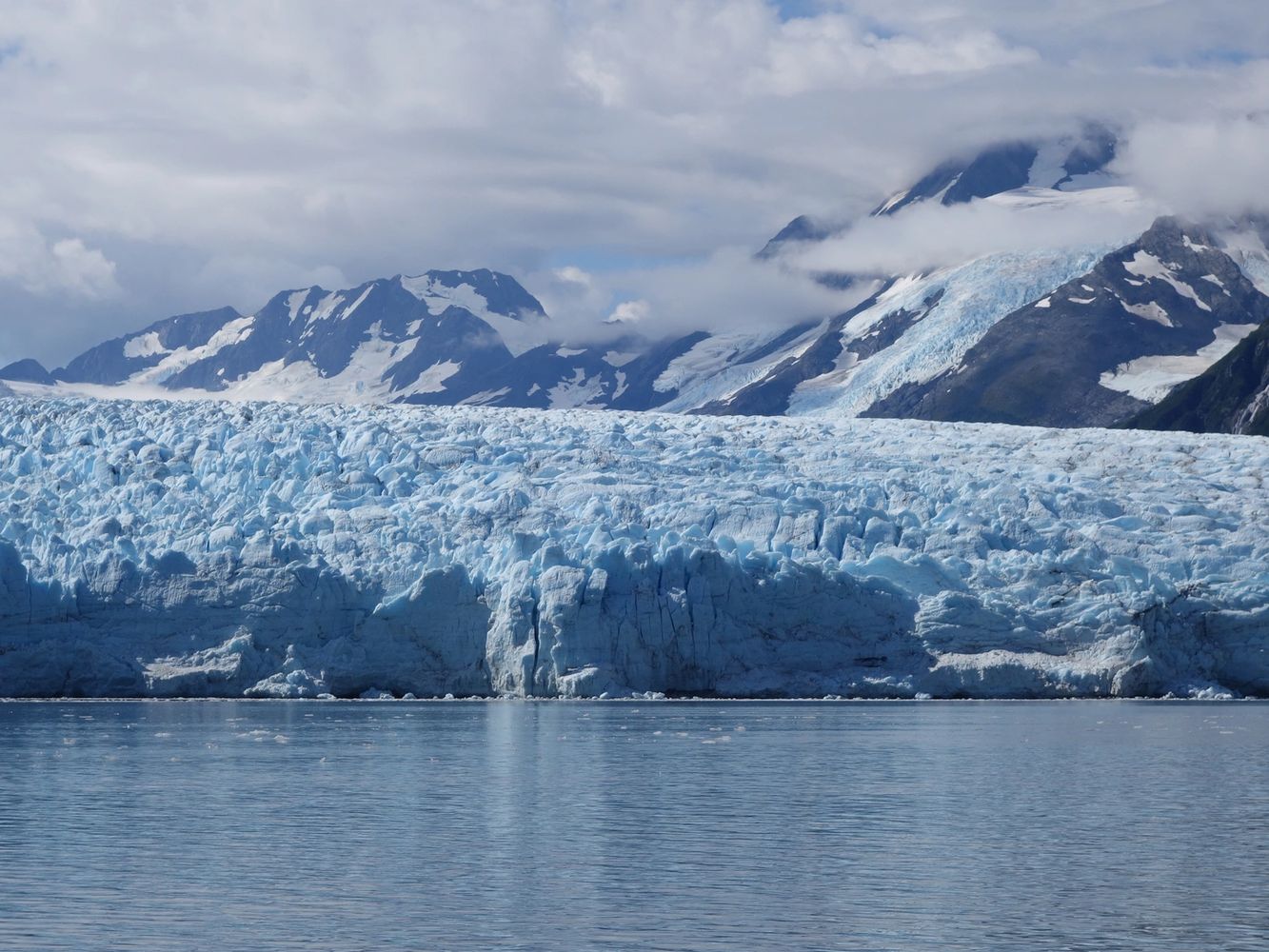 Image shows a glacier against the water, with mountains and clouds behind the glacier.