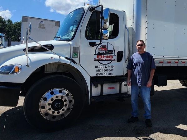 A man stands beside a large white RHS Transport Services truck on a sunny day.