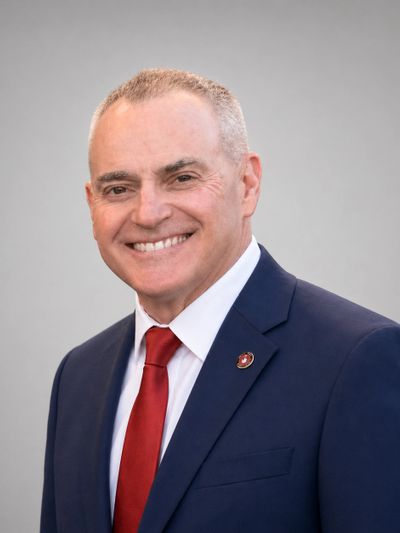 Smiling man in a navy suit with a red tie against a gray background.
