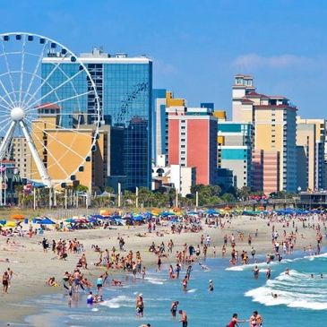 Crowded beach with city skyline and Ferris wheel on a sunny day.
