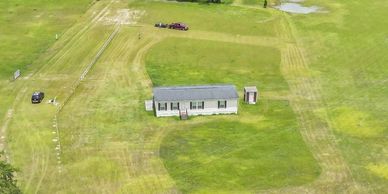 A small house sits isolated in a large grassy field with surrounding trees.