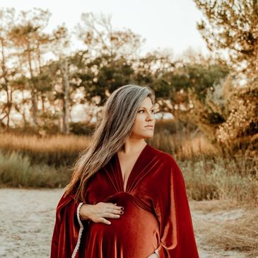 Pregnant woman in a burnt orange velvet dress stands on a beach at sunset