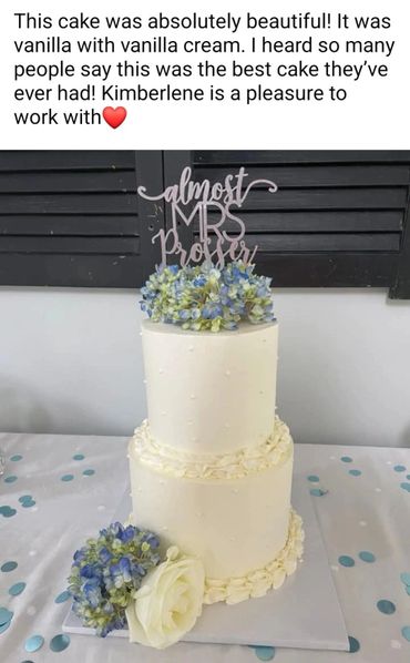 Elegant two-tier vanilla cake with blue and white flowers and a topper saying 'almost MRS Prosser'.