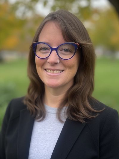 Smiling woman with glasses outdoors in a black blazer and gray shirt.