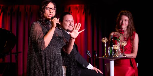Woman speaking on stage with two people seated, one in red dress.