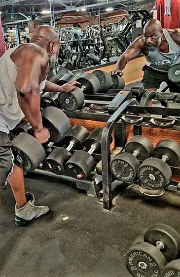 Man lifting dumbbells in gym, reflected in mirror.