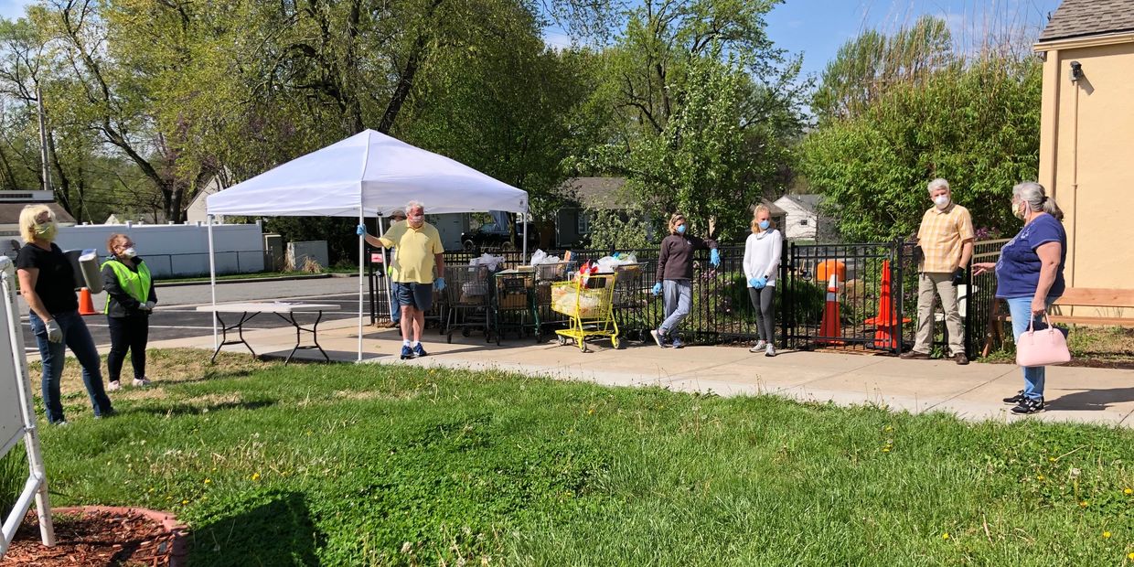 Volunteers wearing masks and gloves stand spaced apart outdoors near a tent and carts.